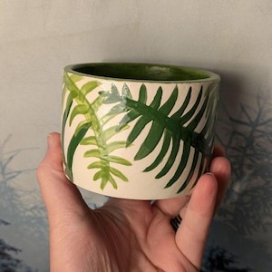 May include: A cylindrical ceramic planter with a cream-colored exterior, adorned with hand-painted green fern leaf designs. The interior of the planter is painted green. The planter is held in a hand.