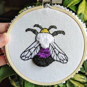 May include: Embroidered hoop art featuring a bee with purple, white, and yellow stripes. The bee is stitched on white fabric and framed in a wooden hoop.