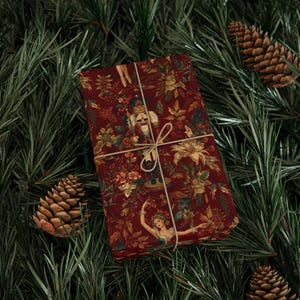 May include: A wrapped gift with a festive design featuring a nutcracker, flowers, and a ballerina on a deep red background. The gift is tied with twine and set against a backdrop of pine needles and pine cones.