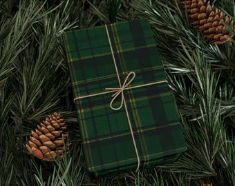 May include: A wrapped gift with green and black plaid wrapping paper, tied with twine. The gift is surrounded by pine needles and pine cones, creating a festive holiday theme.