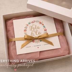 May include: A white gift box containing pink tissue paper and a square card with a floral wreath graphic. The card features the gold text Oh, Lola & Co and the words CREATE • CELEBRATE • CONNECT. A gold ribbon is tied around the card and tissue.