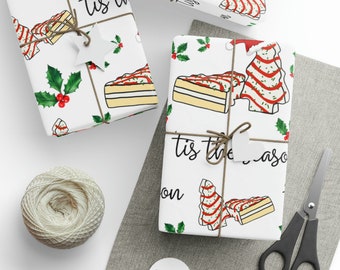 Pode incluir: Papel de embrulho branco com um design festivo com ilustrações de bolo e árvores de Natal, azevinho e a frase "tis the season". Os presentes embrulhados são amarrados com barbante e adornados com etiquetas em forma de estrela.