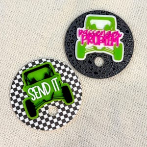 May include: Two round car accessories. One has a black and white checkered pattern with a green off-road vehicle and the words "SEND IT". The other has a dark gray leopard print with a green off-road vehicle and the words "passenger PROBLEM".