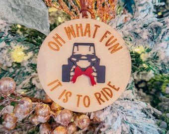 Może przedstawiać: Okrągła drewniana ozdoba choinkowa z napisem "OH WHAT FUN IT IS TO RIDE" w brązowych literach. Ozdoba przedstawia czarno-niebieski wzór pojazdu terenowego z czerwoną kokardą. Ozdoba wisi na choince.