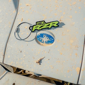 May include: A black and neon green keychain with the text "HELL RZR" on a dirty, light gray surface. The keychain is attached to a metal ring. A blue and white emblem is also visible.