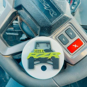May include: A white circular car accessory with the words "HELL RZR" in neon green and a black off-road vehicle graphic. The accessory is on a black steering wheel with gray buttons and a red button.