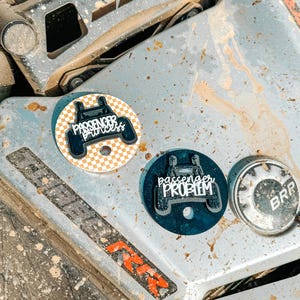 May include: Three round stickers with the text "Passenger Princess" and "Passenger Problem" in white script. One sticker has a checkered background, another is dark blue, and the third has "BRR" on it.