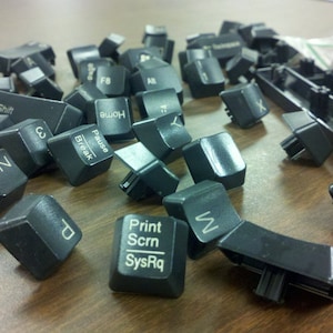May include: A collection of black computer keyboard keys with various letters and symbols printed on them. The keys are scattered on a wooden surface.
