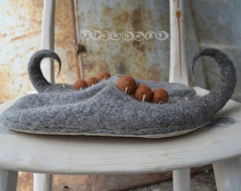 Op de afbeelding: Een paar grijze wollen pantoffels met bruine wollen ballen die erop zijn genaaid. De pantoffels staan op een witte houten stoel.