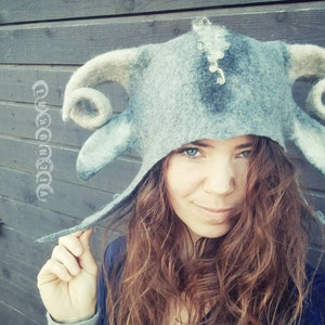 May include: A grey felt hat with curved horns and pointed ears. The hat has a whimsical design, with a light grey and dark grey colour scheme. The hat is worn by a person with long, wavy auburn hair. The background is a dark wooden wall.