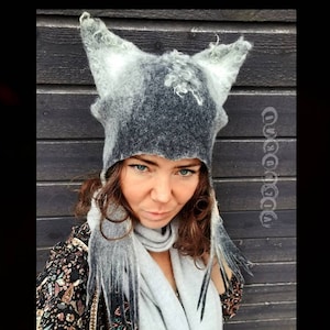 May include: A gray and white felted hat with pointed ears, resembling a wolf or fox, is the focal point. The hat is worn with a matching gray scarf. The person is wearing a floral patterned jacket.