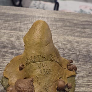 May include: A detailed figurine of a leaf with the text "SOUTH BEND JULY 1983" and the artist's signature. The leaf is a mix of green and brown, with several small acorns around the base. A collectible item.