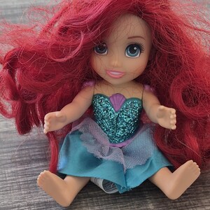 以下が含まれることがあります： 鮮やかな赤毛、青い目、ターコイズと紫色のドレスを着たアリエルの人形。人形は足を伸ばして座っており、笑顔の表情をしています。人形の胴体はキラキラで飾られています。