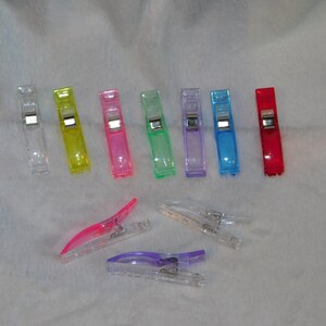 Puede incluir: Una colección de clips de costura coloridos en varios tonos, incluyendo transparente, amarillo, rosa, verde, morado, azul y rojo. Estos clips de plástico están diseñados para proyectos de tela y tienen una bisagra metálica.