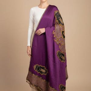 Puede incluir: Un chal de color púrpura intenso con intrincados patrones bordados en oro y negro. El chal tiene un borde decorativo con un borde con flecos. La mujer lleva una camisa blanca de manga larga.
