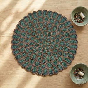 May include: A circular beaded placemat featuring a concentric petal pattern in shades of teal, dark blue, and gold. Two small ceramic bowls are positioned nearby, each containing a collection of colorful glass beads and a small white thread spool.