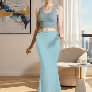 May include: A light blue, floor-length slip skirt with a drawstring waist is shown. The skirt is paired with a light blue tank top. The model is standing in a well-lit room with a painting on the wall and a city view in the background.