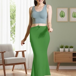 May include: A woman wearing a light blue tank top and a bright green skirt. The skirt is a solid color and has a drawstring at the waist. The woman is standing in front of a window and a chair.
