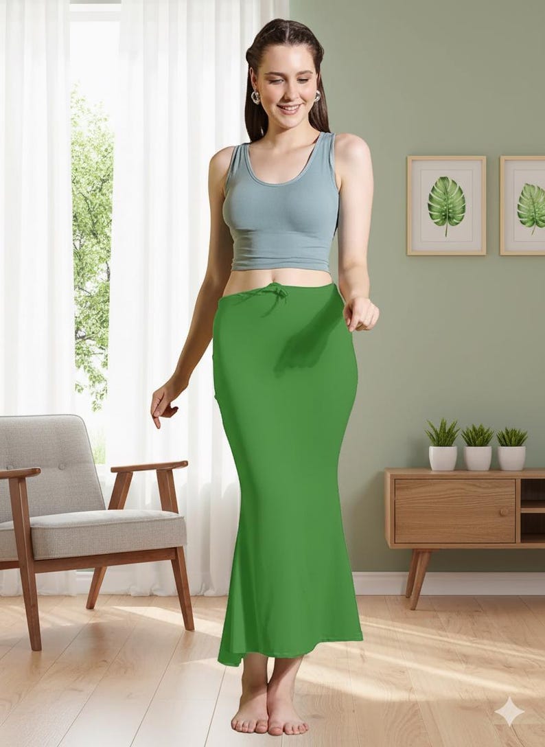 May include: A woman wearing a light blue tank top and a bright green skirt. The skirt is a solid color and has a drawstring at the waist. The woman is standing in front of a window and a chair.