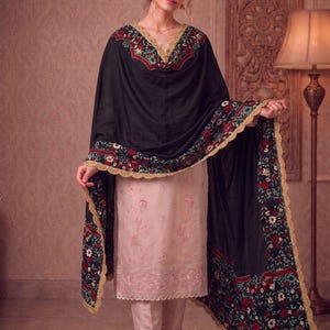 Puede incluir: Una túnica de color rosa claro hasta la rodilla con pantalones rectos a juego, combinada con un gran chal negro con un vibrante estampado floral en rojo, blanco y verde, acabado con un borde de encaje dorado festoneado en todo el contorno.