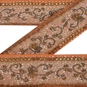 Może przedstawiać: Zbliżenie dekoracyjnego wykończenia w odcieniach pomarańczu i brązu. Wykończenie charakteryzuje się skomplikowanymi haftami, cekinami i koralikami. Wzór obejmuje wzory kwiatowe i zwojowe, z obramowaniem w kształcie łezki.