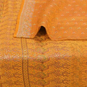 May include: Mustard yellow textile with intricate floral and paisley patterns. The fabric is folded, revealing a glimpse of the underside. A detailed border with floral motifs frames the edge. The overall design suggests a decorative textile, possibly for home decor.