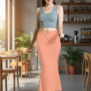 May include: A woman wearing a light blue tank top and a peach-colored, floor-length skirt. The skirt has a drawstring waist. The background shows a cafe setting with wooden tables, chairs, and shelves.