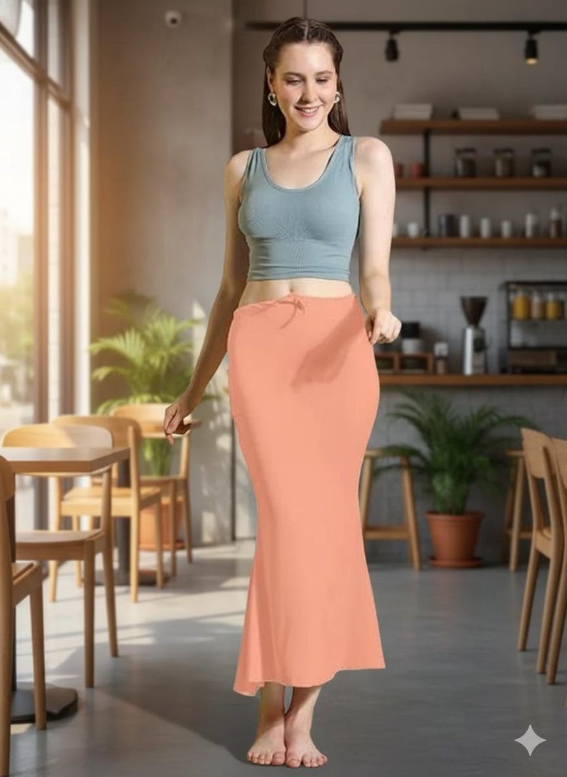 May include: A woman wearing a light blue tank top and a peach-colored, floor-length skirt. The skirt has a drawstring waist. The background shows a cafe setting with wooden tables, chairs, and shelves.
