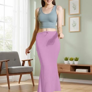 May include: A woman wearing a light blue tank top and a long, light purple skirt. The skirt has a drawstring waist and a fitted silhouette. The background includes a wooden floor, a gray chair, and a wooden cabinet with plants.
