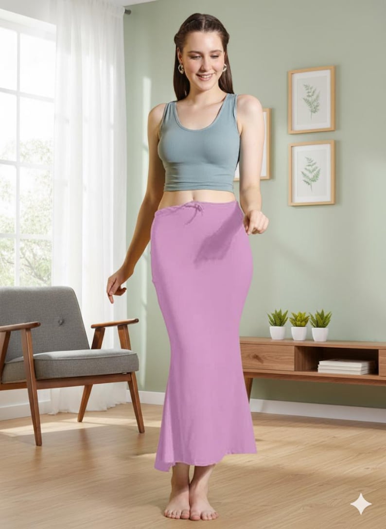 May include: A woman wearing a light blue tank top and a long, light purple skirt. The skirt has a drawstring waist and a fitted silhouette. The background includes a wooden floor, a gray chair, and a wooden cabinet with plants.
