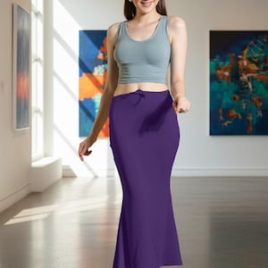 May include: A woman wearing a light gray crop top and a long, deep purple skirt. The skirt has a slight flare at the bottom. The woman is in a gallery setting with colorful abstract paintings on the walls.