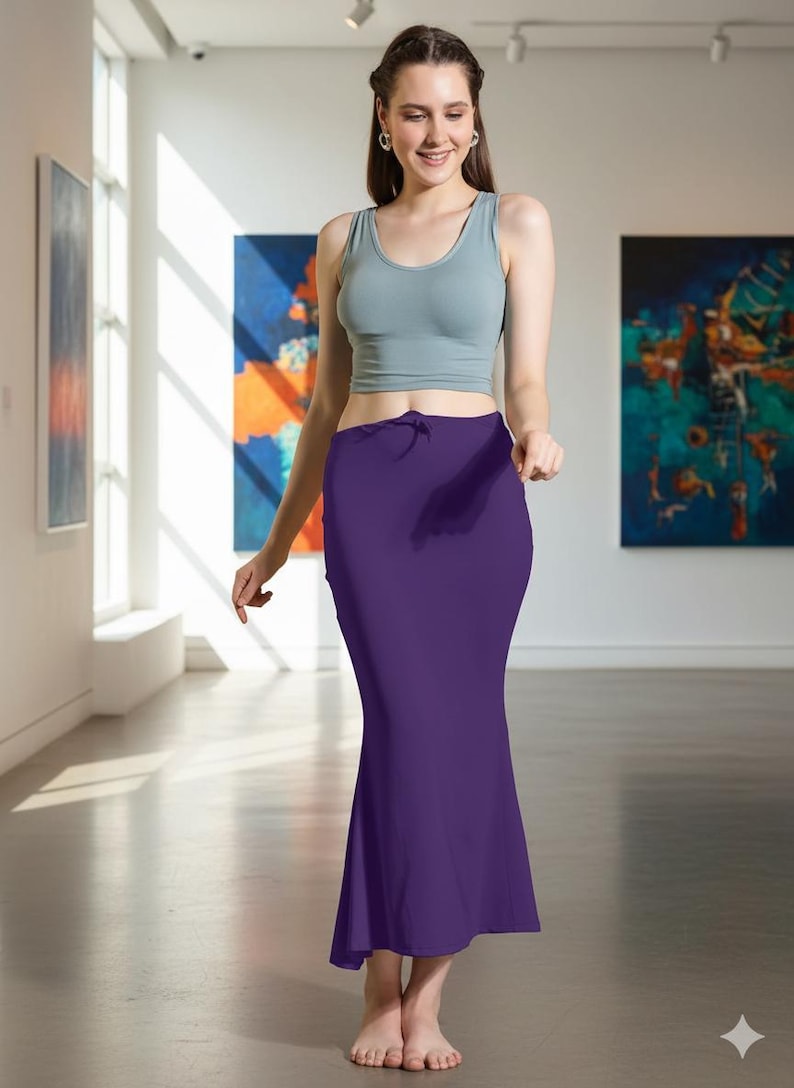 May include: A woman wearing a light gray crop top and a long, deep purple skirt. The skirt has a slight flare at the bottom. The woman is in a gallery setting with colorful abstract paintings on the walls.