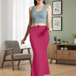 May include: A magenta-colored, floor-length skirt with a slight flare at the bottom. The skirt has a drawstring waist and is paired with a light blue tank top. The background includes a wooden cabinet, framed botanical prints, and a gray armchair.