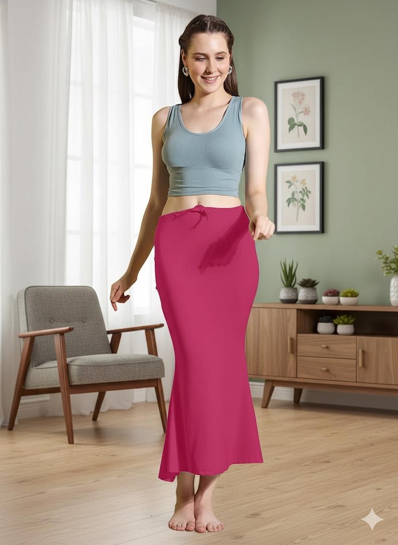 May include: A magenta-colored, floor-length skirt with a slight flare at the bottom. The skirt has a drawstring waist and is paired with a light blue tank top. The background includes a wooden cabinet, framed botanical prints, and a gray armchair.