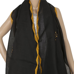 May include: A black scarf featuring a mustard yellow border with a repeating triangular pattern. The scarf is draped over the shoulders and displays subtle yellow embroidered details on the fabric, showcasing a lightweight and semi-sheer texture.