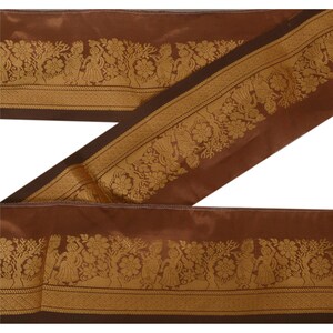 May include: Brown fabric ribbon with a gold floral and figure design. The ribbon has a dark brown border and a repeating pattern of figures, trees, and flowers. The ribbon is folded over itself.