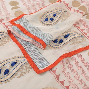 May include: A sheer off-white fabric featuring embroidered paisley motifs in gold and blue thread. The textile includes a vibrant orange border with a chevron pattern and red printed spiral designs, showcasing intricate needlework and layered textures.