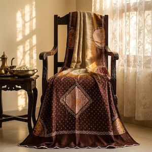 May include: A brown and tan textile draped over a wooden chair. The fabric features a central diamond pattern, intricate small white dot motifs, and larger semi-circular designs along the edges. The material has a smooth, flowing appearance.