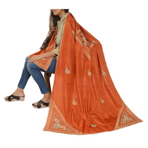 May include: An orange shawl with embroidered floral designs and a decorative border. The shawl is draped over a person wearing a green shirt and blue jeans. The shawl's fabric has a slight sheen.