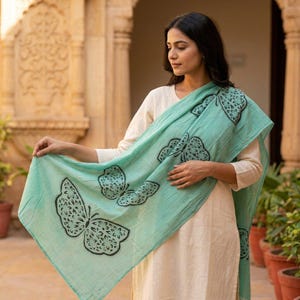 May include: A light turquoise scarf featuring a repeating pattern of black butterfly silhouettes with intricate floral-like details within the wings. The scarf is draped over a plain, long-sleeved, off-white garment with a simple round neckline.