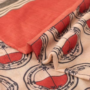 May include: A textile fabric featuring a repeating pattern of red circular motifs outlined in black on a beige background. The fabric includes a solid red section and a border with thin black and white stripes, showing a soft, folded texture.