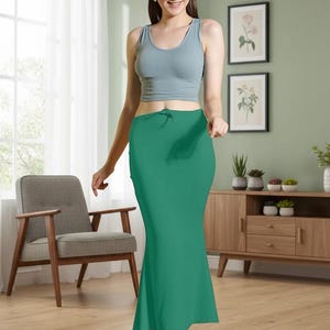 May include: A woman wearing a light blue tank top and a long, emerald green skirt. The skirt has a fitted silhouette that flares out slightly at the bottom. The woman is standing in a room with a chair, a cabinet, and framed artwork.