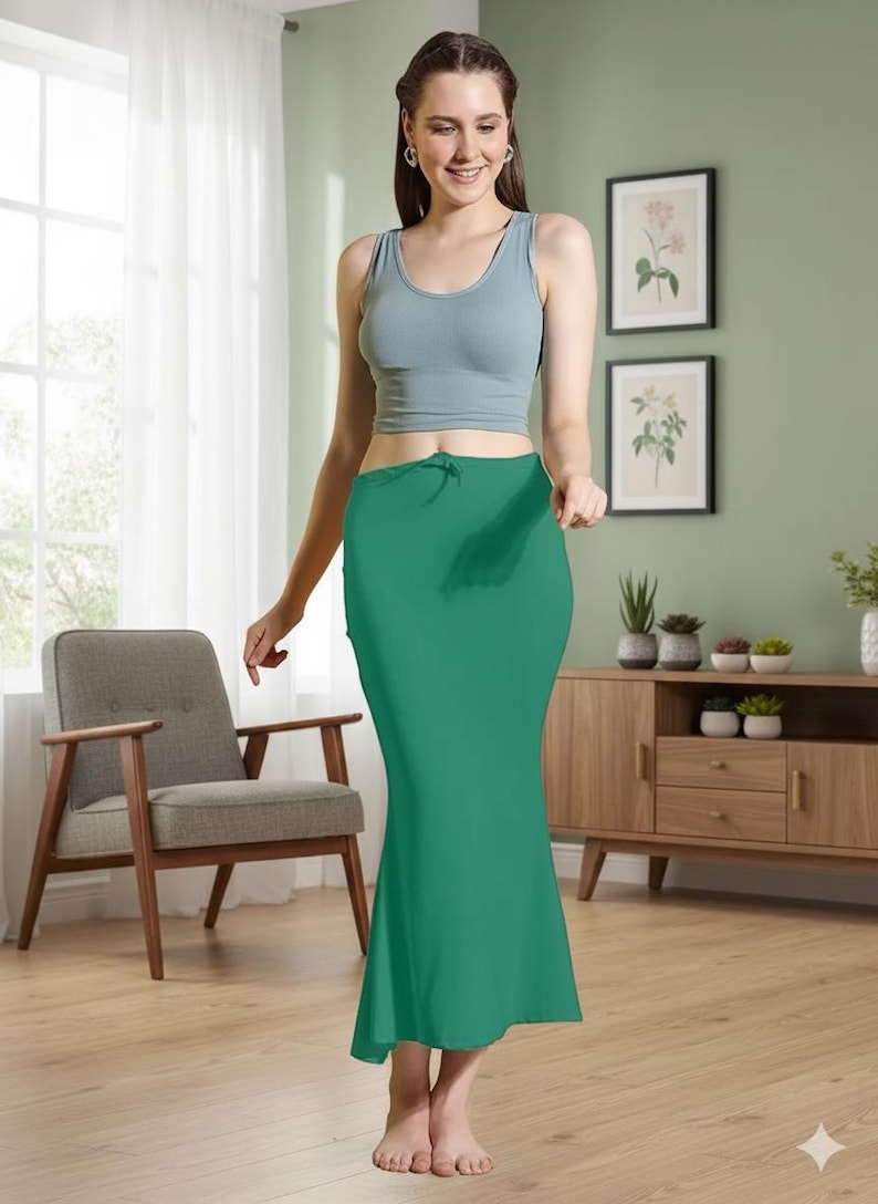 May include: A woman wearing a light blue tank top and a long, emerald green skirt. The skirt has a fitted silhouette that flares out slightly at the bottom. The woman is standing in a room with a chair, a cabinet, and framed artwork.