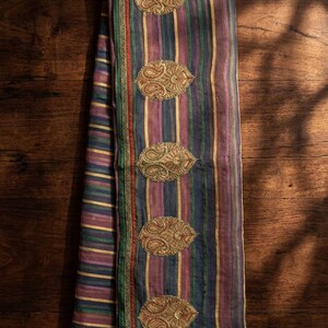 May include: A folded fabric textile featuring vertical stripes in shades of purple, green, and gold. The edge is finished with a green border and a thin red line. Five oval-shaped gold embroidered patches with intricate paisley patterns are aligned.