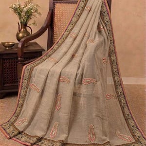 Può includere: Un sari beige caratterizzato da un sottile motivo a griglia e ricami paisley rosa e oro sparsi. Il capo è rifinito con un'ampia bordura scura decorata con intricati motivi floreali dorati e un sottile bordo rosa lungo i margini.
