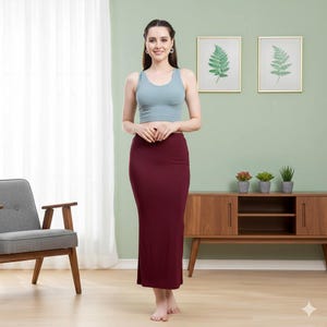 May include: A woman wearing a light blue crop top and a burgundy pencil skirt. The skirt is long, reaching the ankles. The woman is standing in front of a light green wall with framed botanical prints and a wooden cabinet.