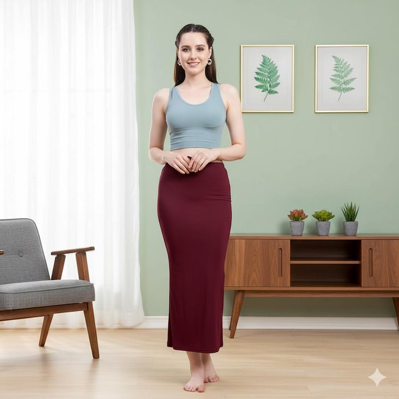 May include: A woman wearing a light blue crop top and a burgundy pencil skirt. The skirt is long, reaching the ankles. The woman is standing in front of a light green wall with framed botanical prints and a wooden cabinet.