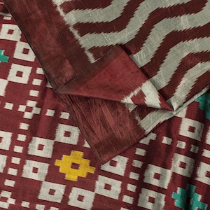 May include: A textile fabric featuring a deep maroon base with a geometric pattern of white squares and zigzags. The design includes small yellow and teal accents integrated into the grid, showcasing a woven texture with visible thread details.