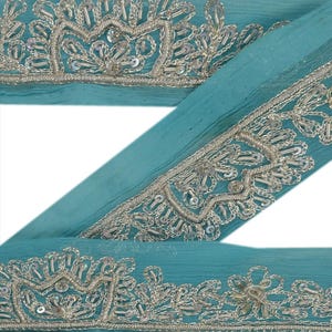 May include: Teal blue fabric trim with silver embroidery and sequins. The trim features a repeating pattern of floral designs.