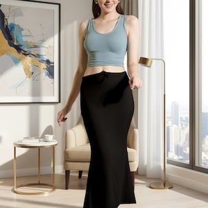 May include: A woman wearing a light blue tank top and a black, floor-length skirt. The skirt has a slight flare at the bottom. The woman is standing in a well-lit room with a window and a gold lamp.